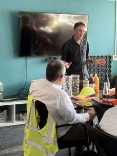 Man presenting to seated group in meeting room with TV, discussing rigid plastics recycling.